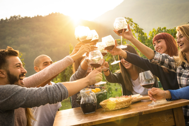 Happy friends having fun outdoors – Young people enjoying harvest time together at farmhouse vineyard countryside – Youth and friendship concept – Focus on hands toasting wine glasses with sun flare